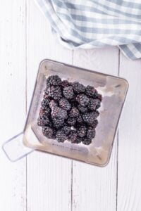 Blackberries in a blender on a white wooden surface with a checkered cloth nearby.
