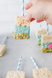 A hand holds a rice crispy treat on a blue-striped stick dipped in blue coating.