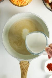 A hand pours milk from a measuring cup into a white saucepan on a stovetop.