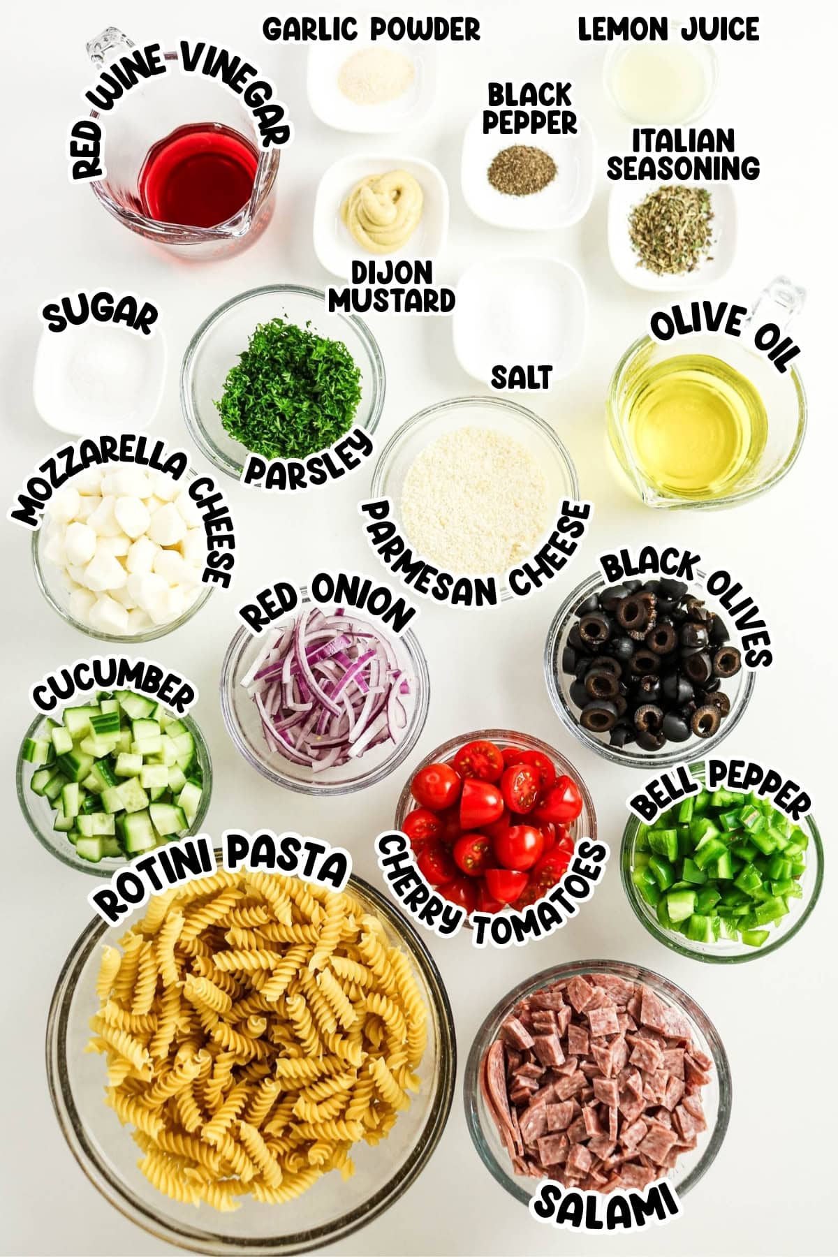 Labeled ingredients for pasta salad arranged on a white surface in small bowls.