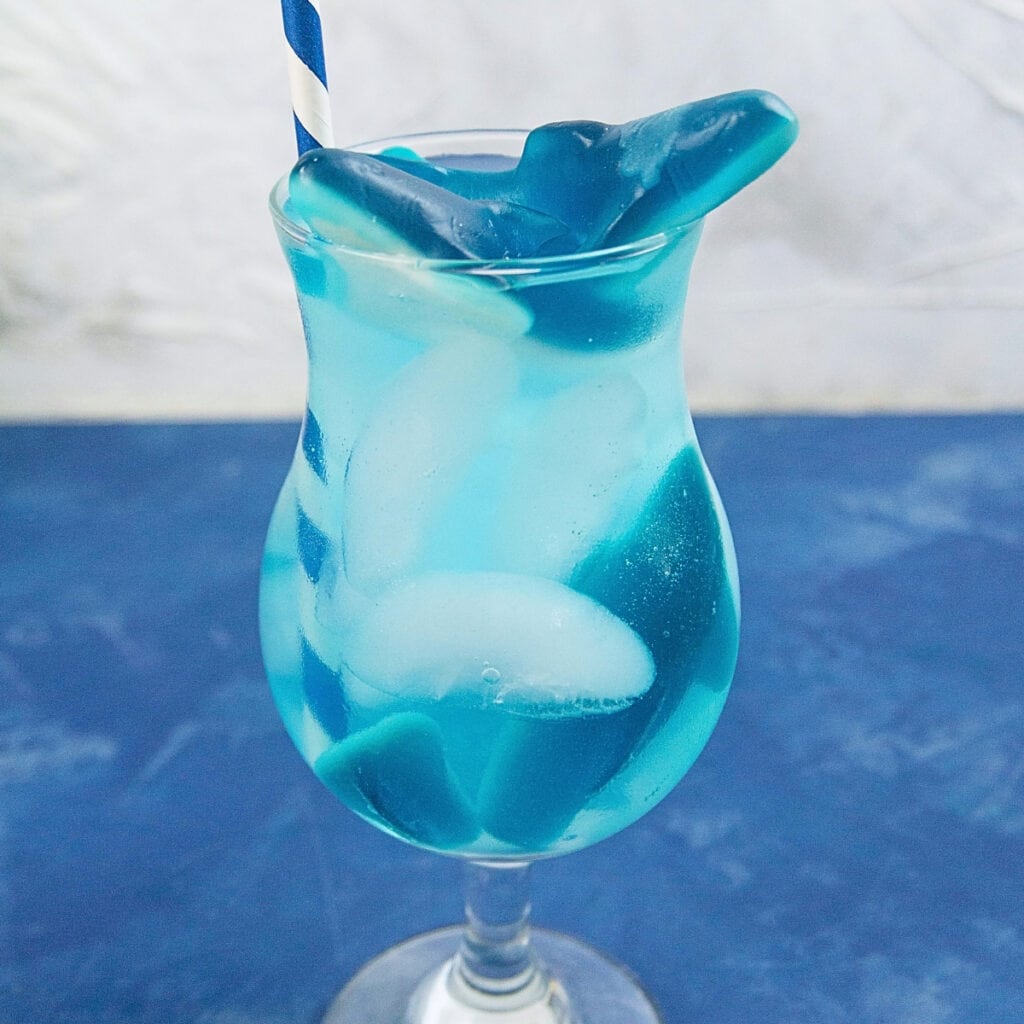 A blue drink with ice, gummy sharks, and a striped straw in a cocktail glass.