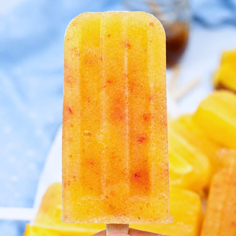 A hand holds a yellow-orange fruit popsicle with red specks.