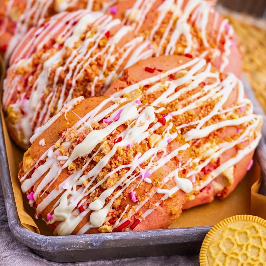 Pink cookies with white drizzle, sprinkles, and cookie crumbs on a baking tray.