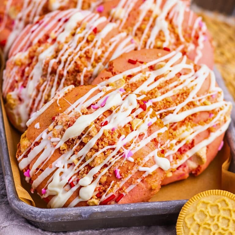 Pink cookies with white drizzle, sprinkles, and cookie crumbs on a baking tray.