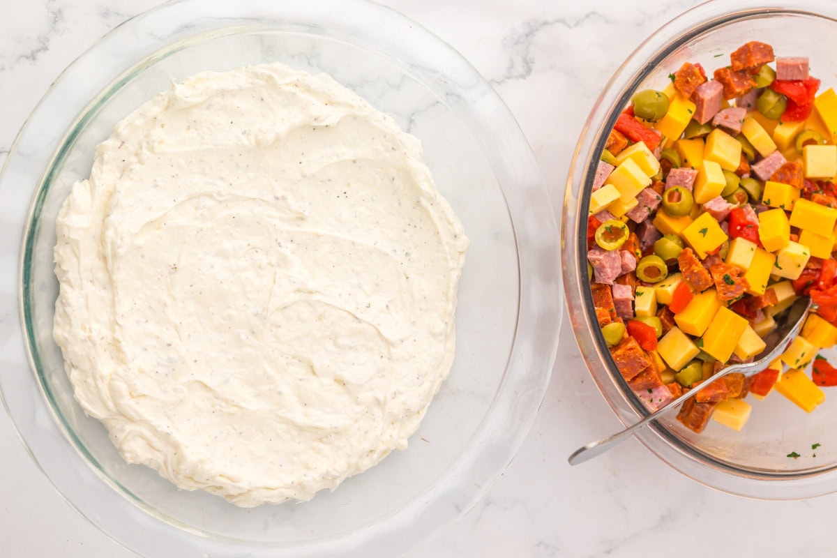 A glass dish with creamy dip and a bowl of chopped meats, cheese, and veggies.