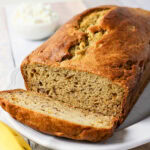 A loaf of banana bread with one slice cut, on a white plate.