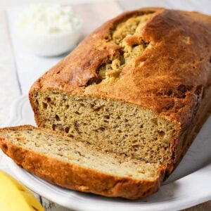 A loaf of banana bread with one slice cut, on a white plate.