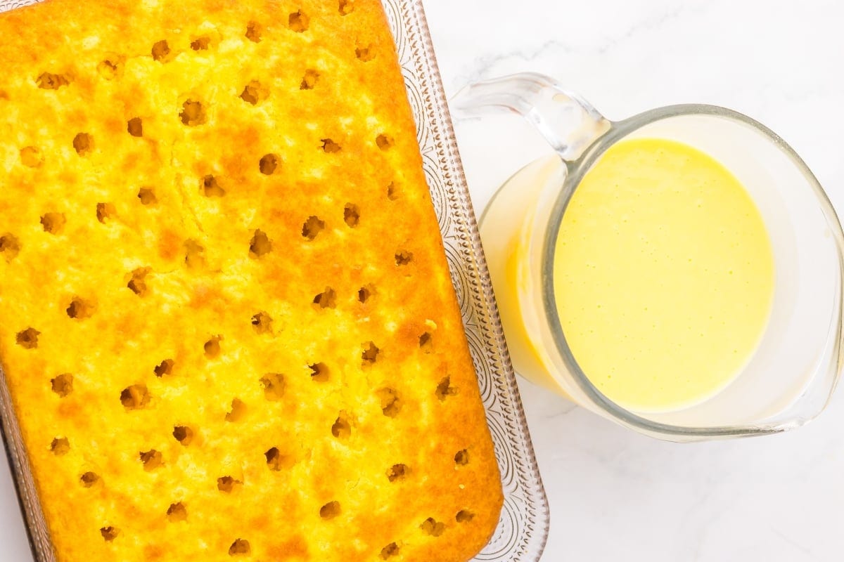 Golden cake with holes next to a glass pitcher of yellow sauce on a white surface.