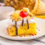 Slice of yellow cake with whipped topping, pineapple, cherry, and chocolate drizzle.