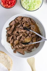 Cooked beef strips in a skillet with tongs, surrounded by lettuce, tomato, and bread.