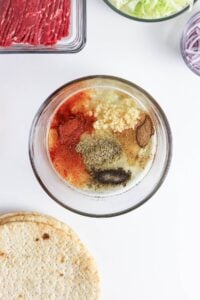 A bowl of assorted spices and minced garlic, surrounded by sandwich ingredients.