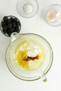 Glass bowl with baking ingredients, blackberries, flour, and eggshells on the side.