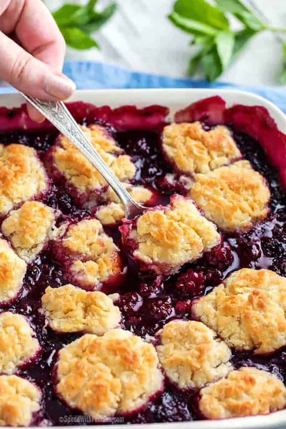 A hand scoops blackberry cobbler with a spoon from a baking dish.