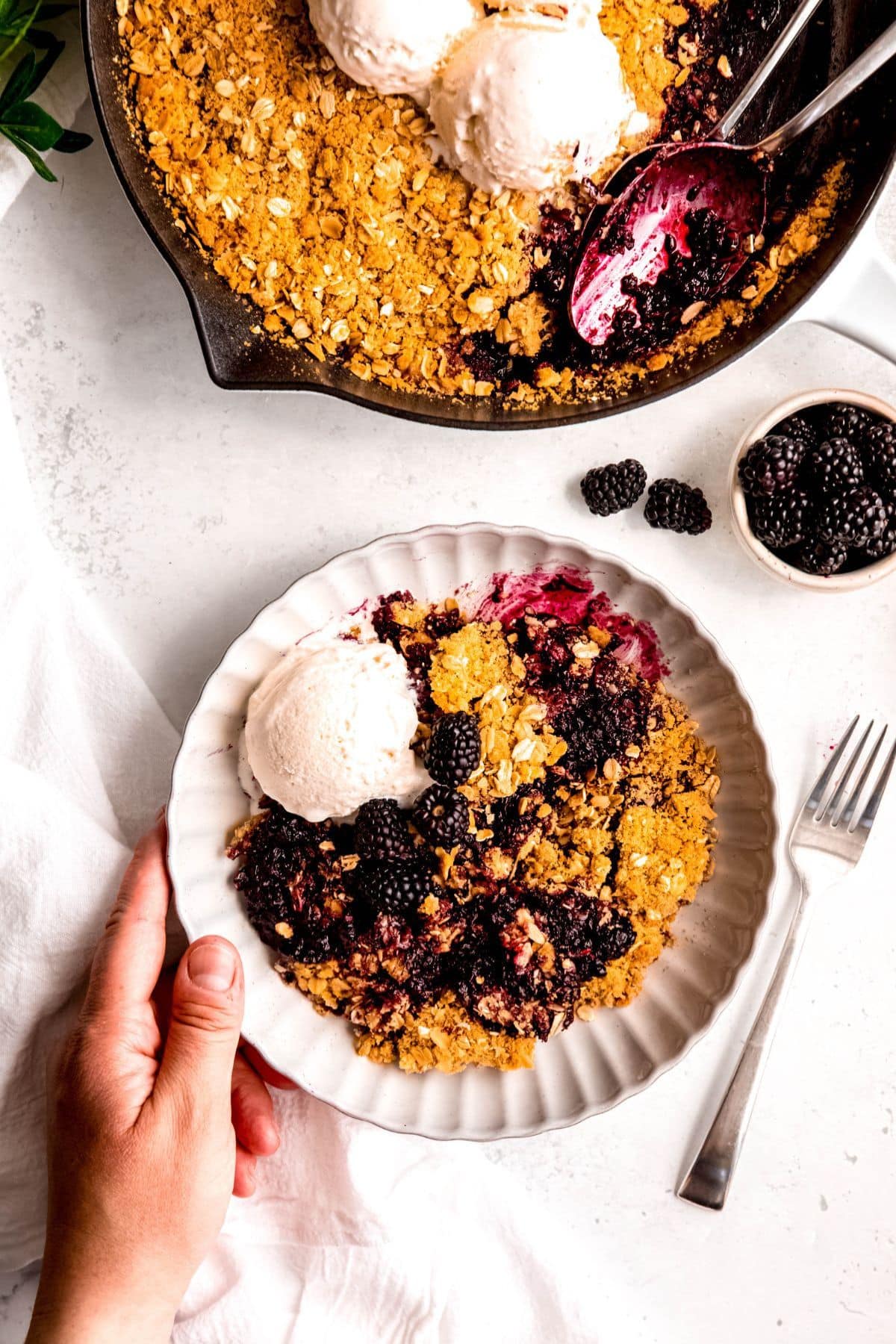 A plate of blackberry crisp with ice cream, beside a pan and a small bowl of berries.