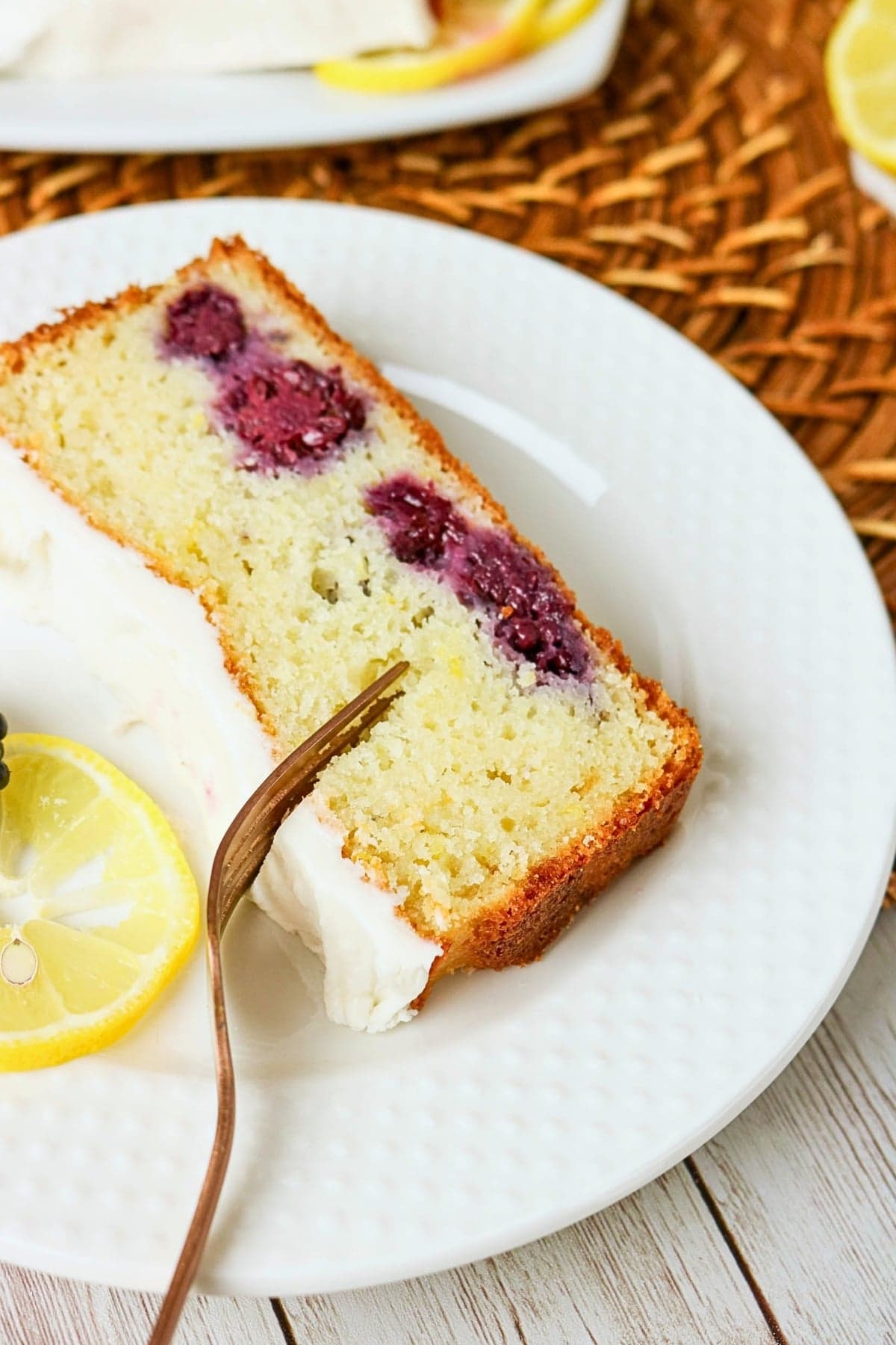 A slice of berry cake with white icing and a lemon slice on a white plate.