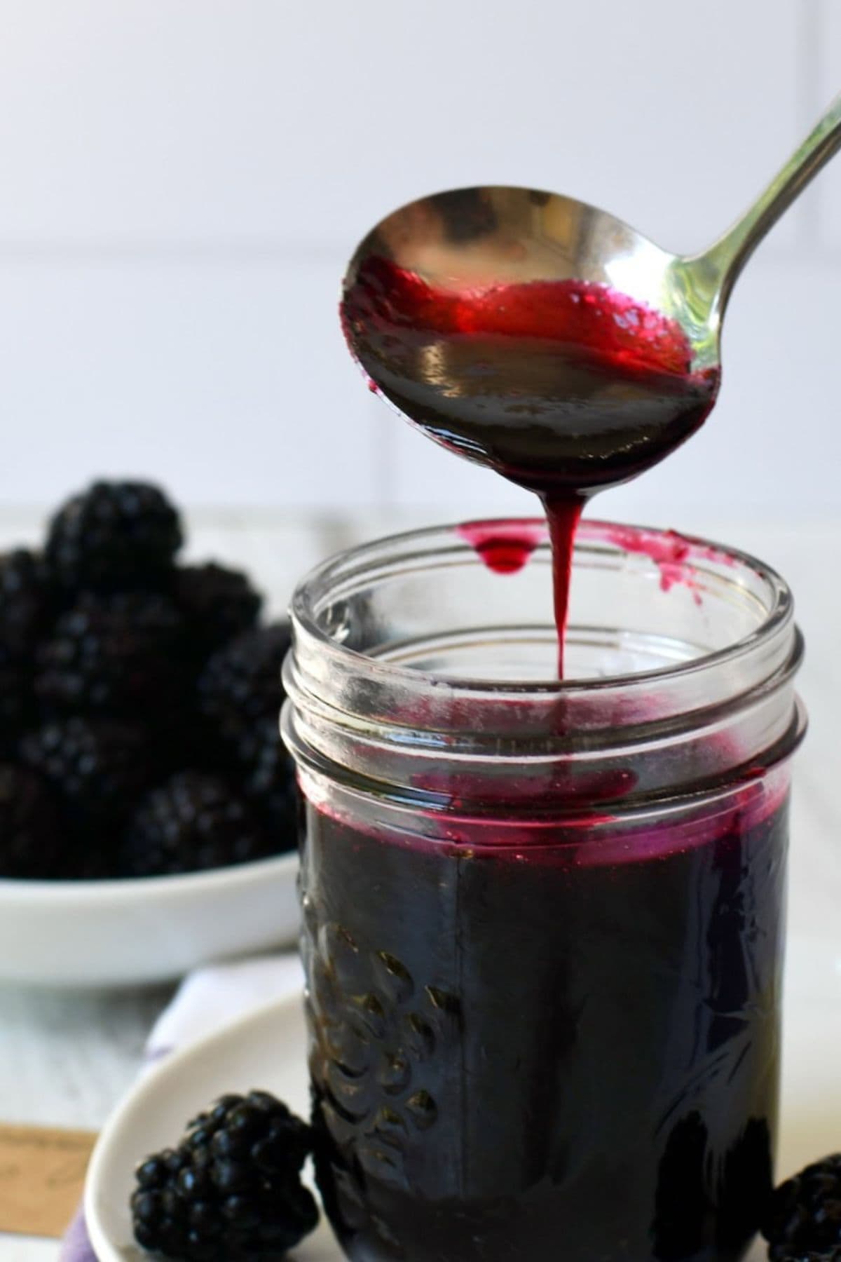 A spoon drizzles blackberry syrup into a glass jar with blackberries nearby.