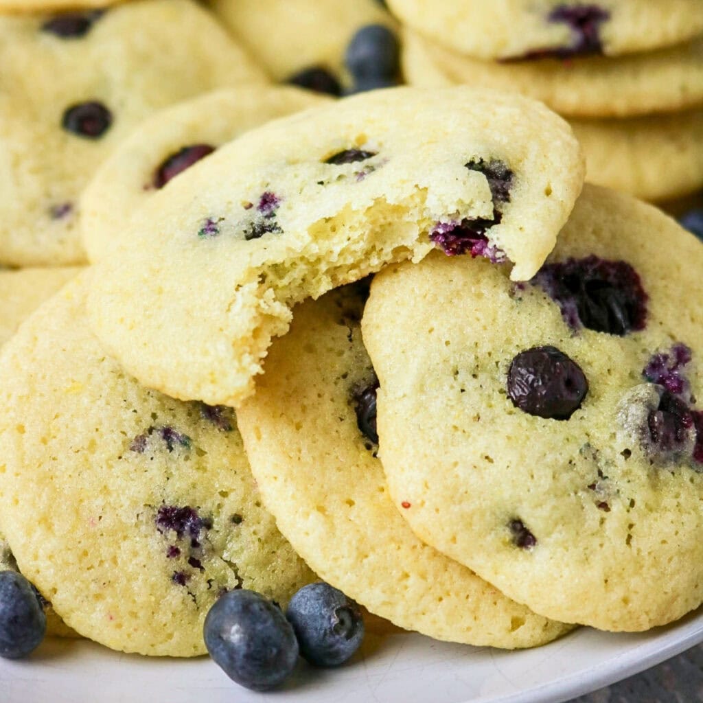 A plate of blueberry cookies, one with a bite taken out, and fresh blueberries.