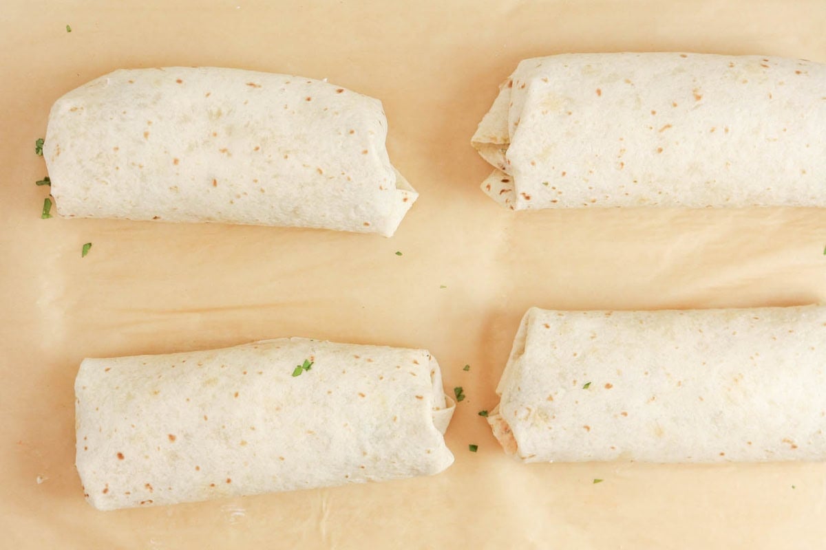 Four rolled tortillas on parchment paper, sprinkled with small bits of herbs.