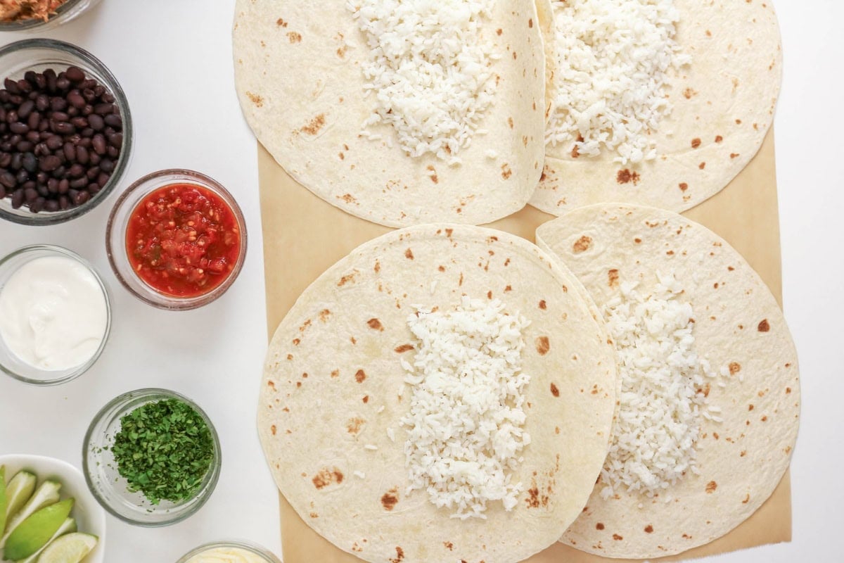 Four tortillas topped with white rice, surrounded by bowls of various taco ingredients.