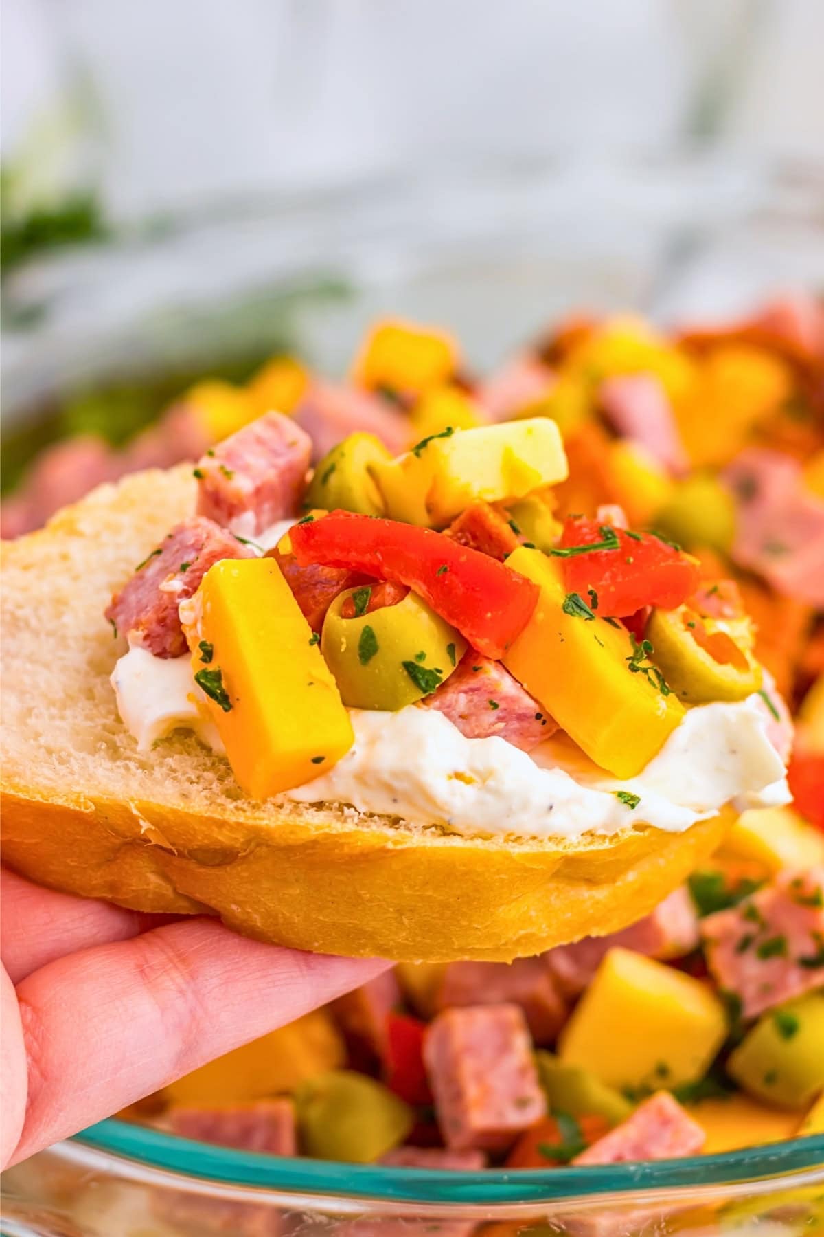 A hand holds bread topped with creamy spread, cheese, olives, meat, and peppers.