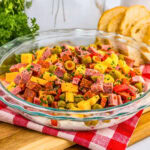 A glass dish of chopped cheese, salami, olives, and herbs, with bread slices nearby.