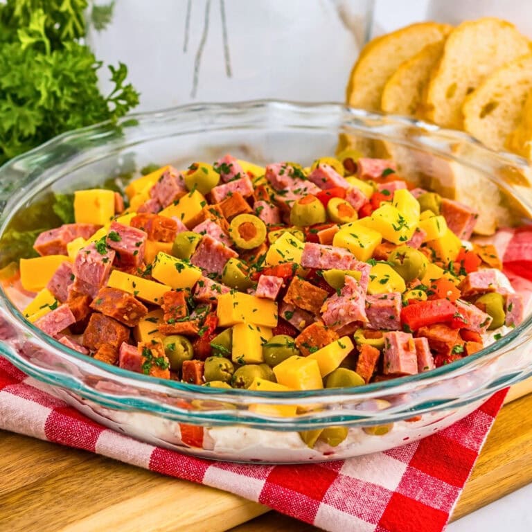 A glass dish of chopped cheese, salami, olives, and herbs, with bread slices nearby.