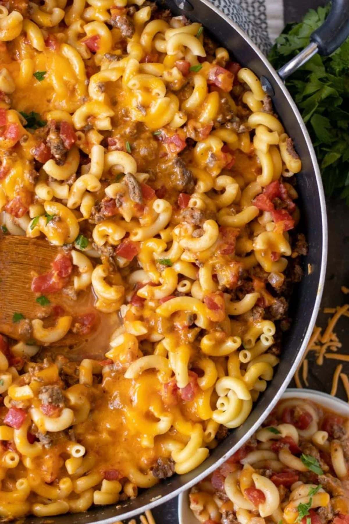 A pan of cheesy macaroni with ground beef and tomatoes, being stirred with a spoon.