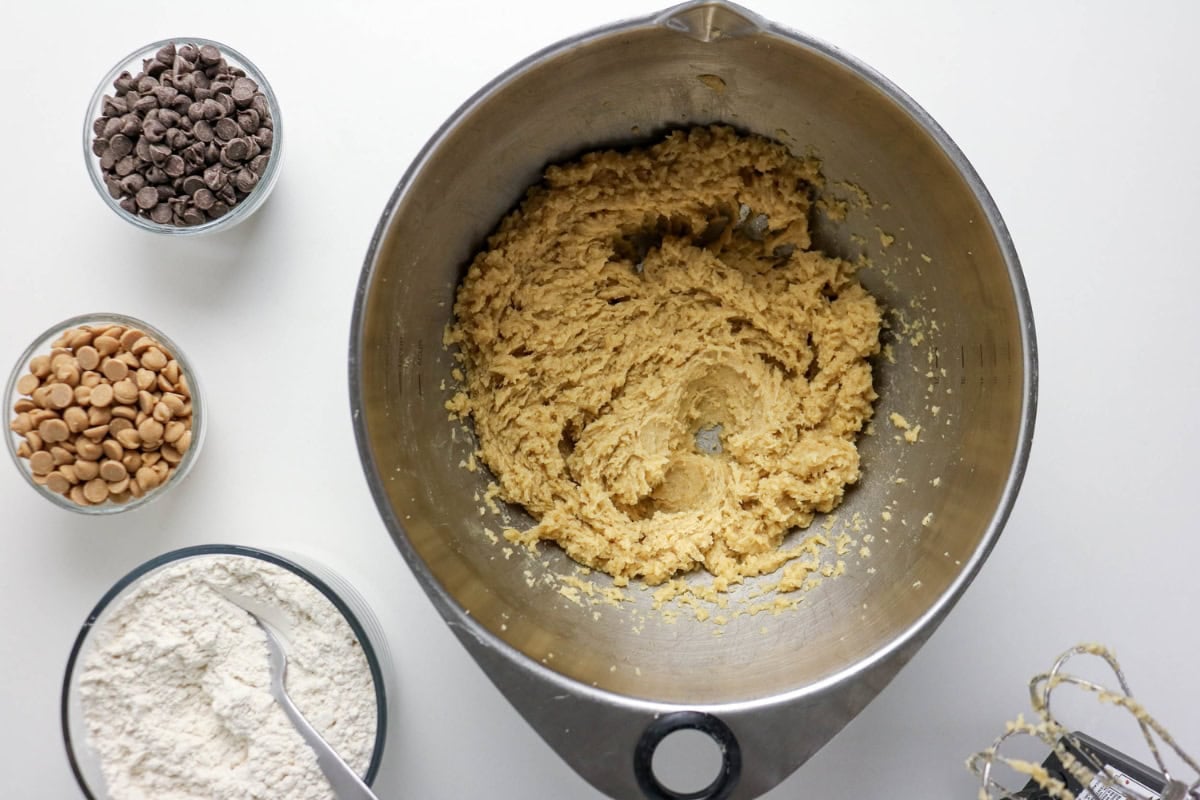 A mixing bowl with cookie dough, flour, chocolate, and peanut butter chips nearby.