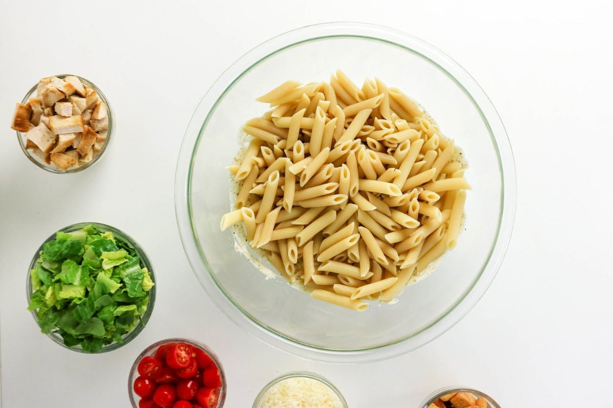 Bowl of penne pasta surrounded by small bowls of salad, tomatoes, and toppings.