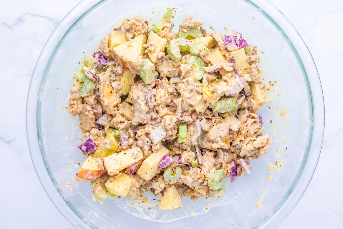 Chicken salad with apples, celery, and nuts in a glass bowl on a white surface.