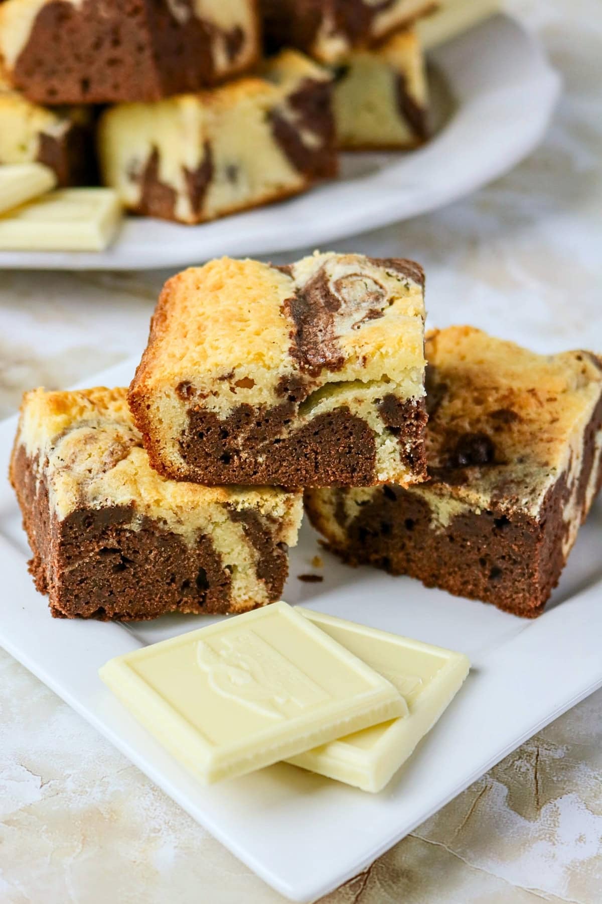 Marble cheesecake brownies stacked on a plate with pieces of white chocolate.