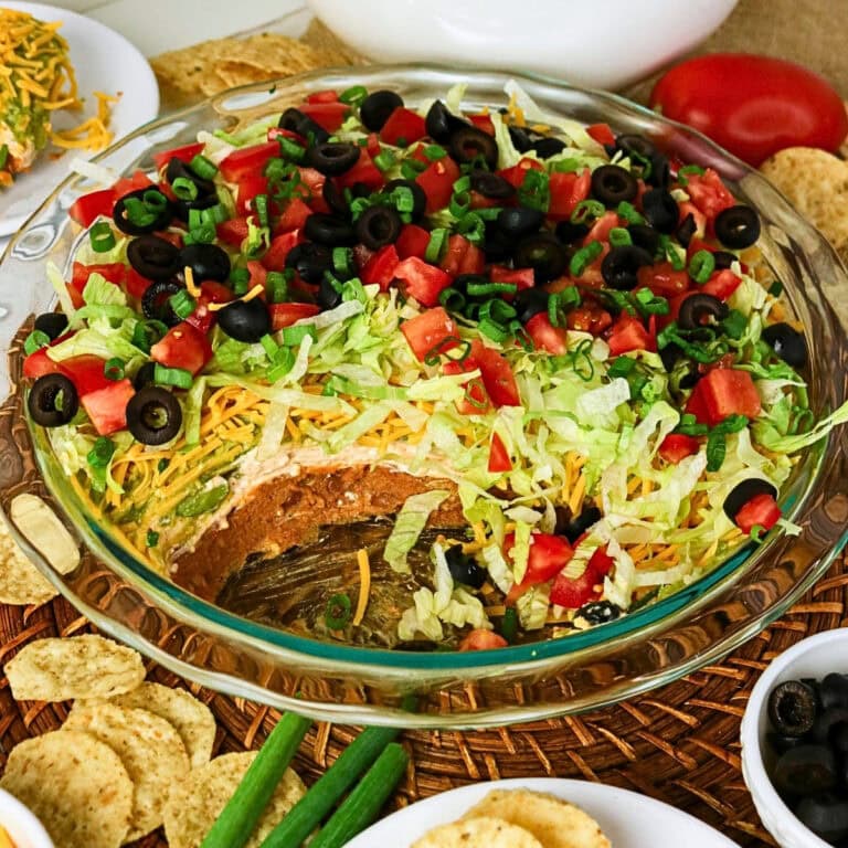 A glass dish of layered taco dip topped with lettuce, tomatoes, olives, and cheese.