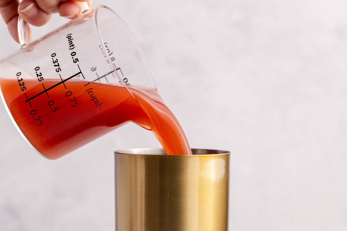A hand pours red liquid from a measuring cup into a metal container.