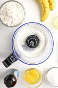 Food processor with batter, eggs, bananas, flour, sugar, and vanilla on counter.