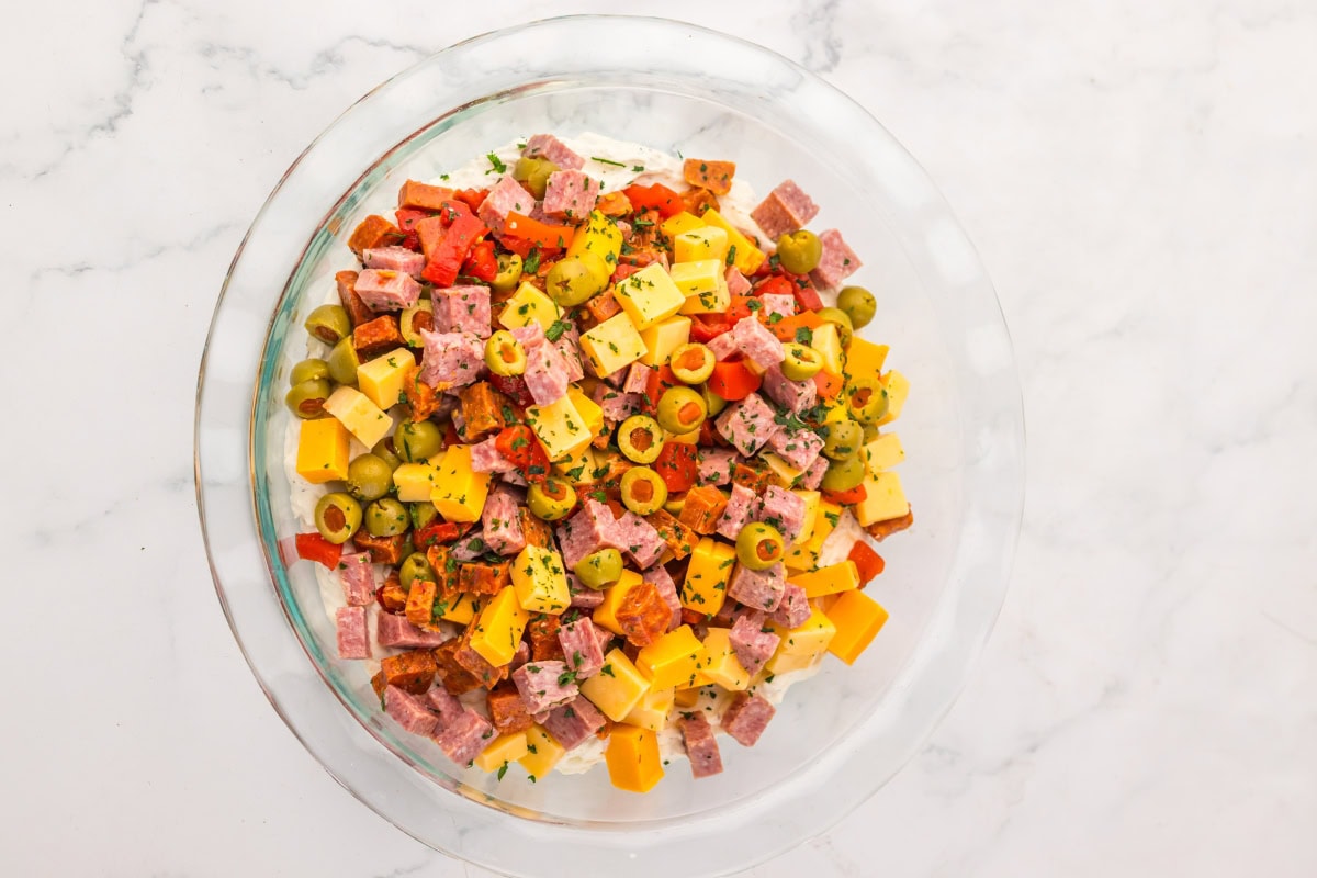 A glass bowl filled with cubed cheese, salami, olives, and red peppers.