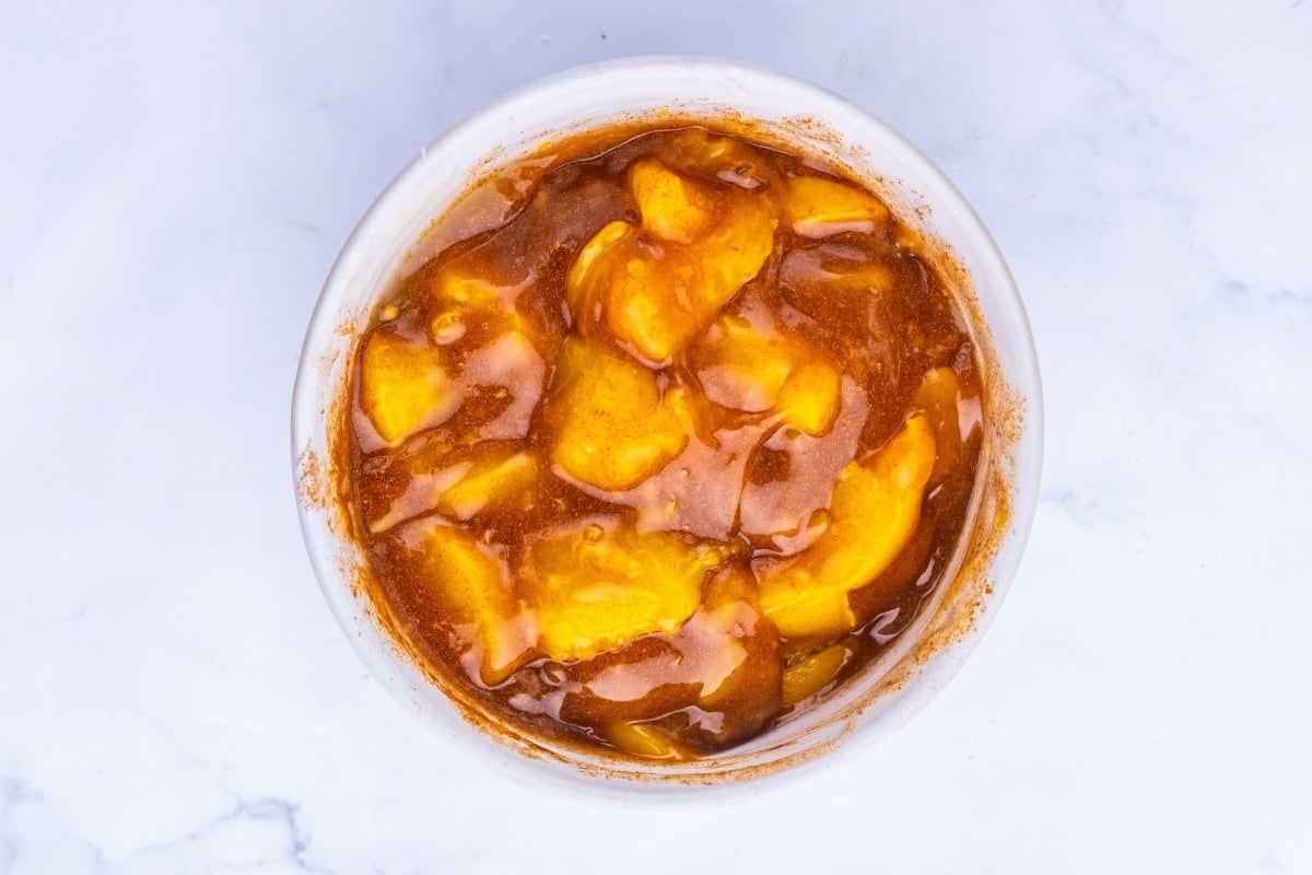 A bowl of sliced peaches mixed with cinnamon and syrup on a white surface.
