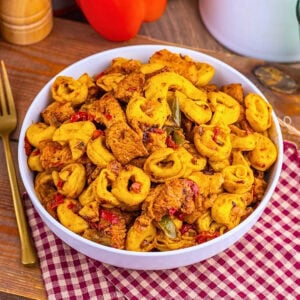 A bowl of tortellini pasta with chicken and vegetables on a red checkered cloth.
