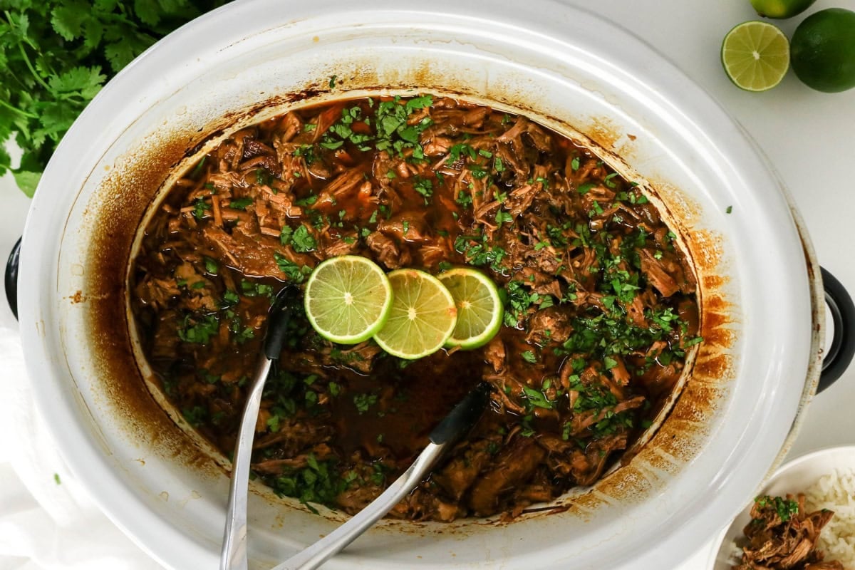 Shredded beef in a slow cooker, garnished with lime slices and fresh cilantro.