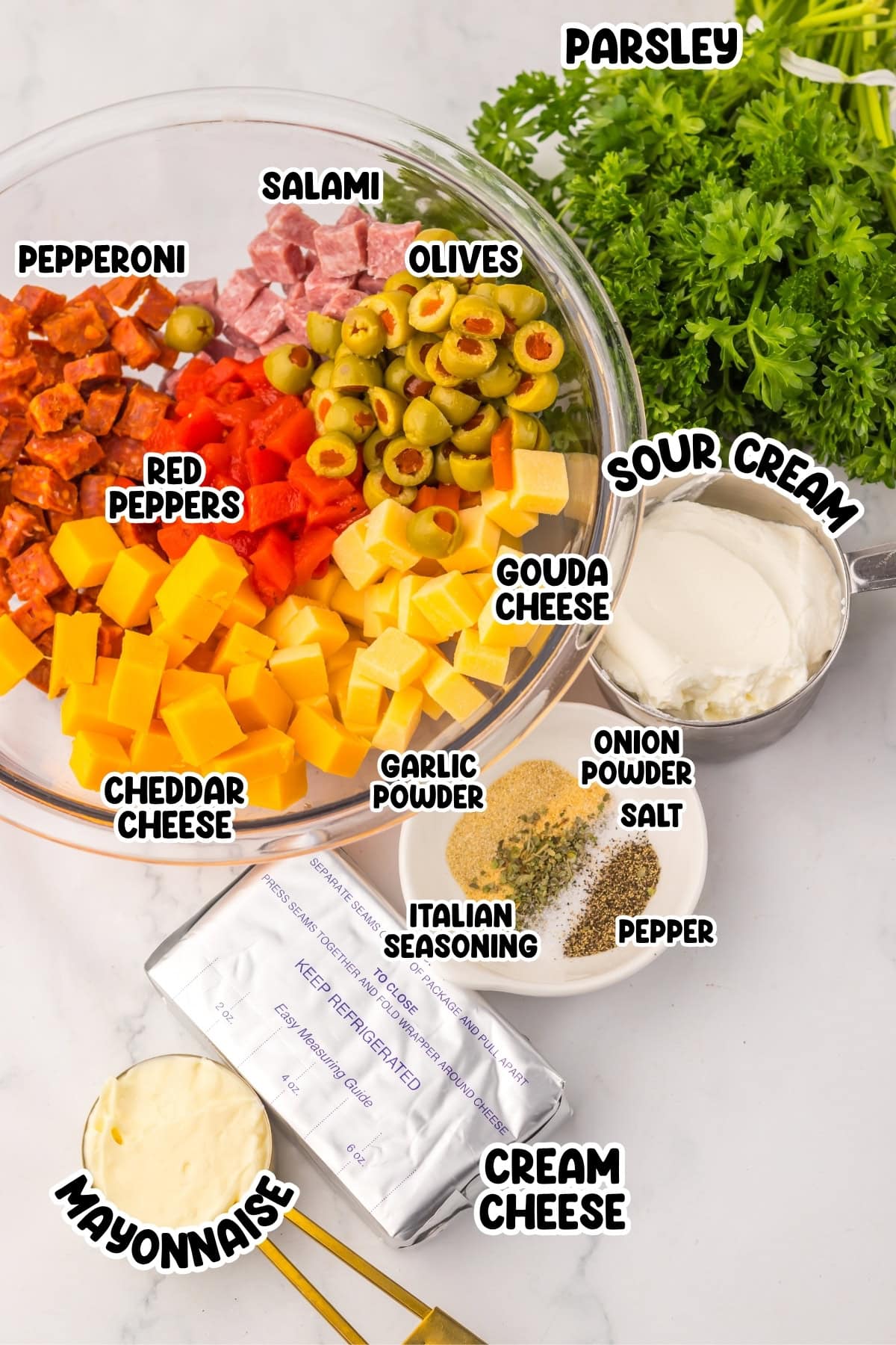 Bowl of diced cheese, meats, olives, peppers, with herbs and dairy ingredients nearby.