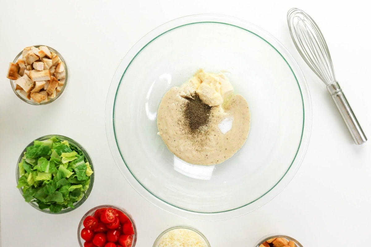 Glass bowl with dressing ingredients surrounded by salad fixings and a whisk.