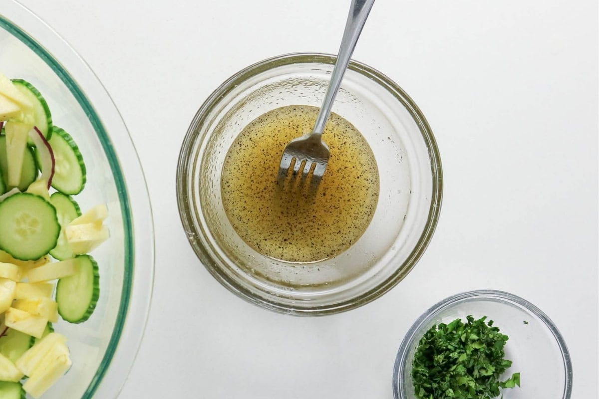 Bowl of vinaigrette with a fork, next to sliced veggies and chopped herbs.