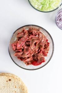 Sliced raw beef with seasoning in a glass bowl, surrounded by pita and veggies.