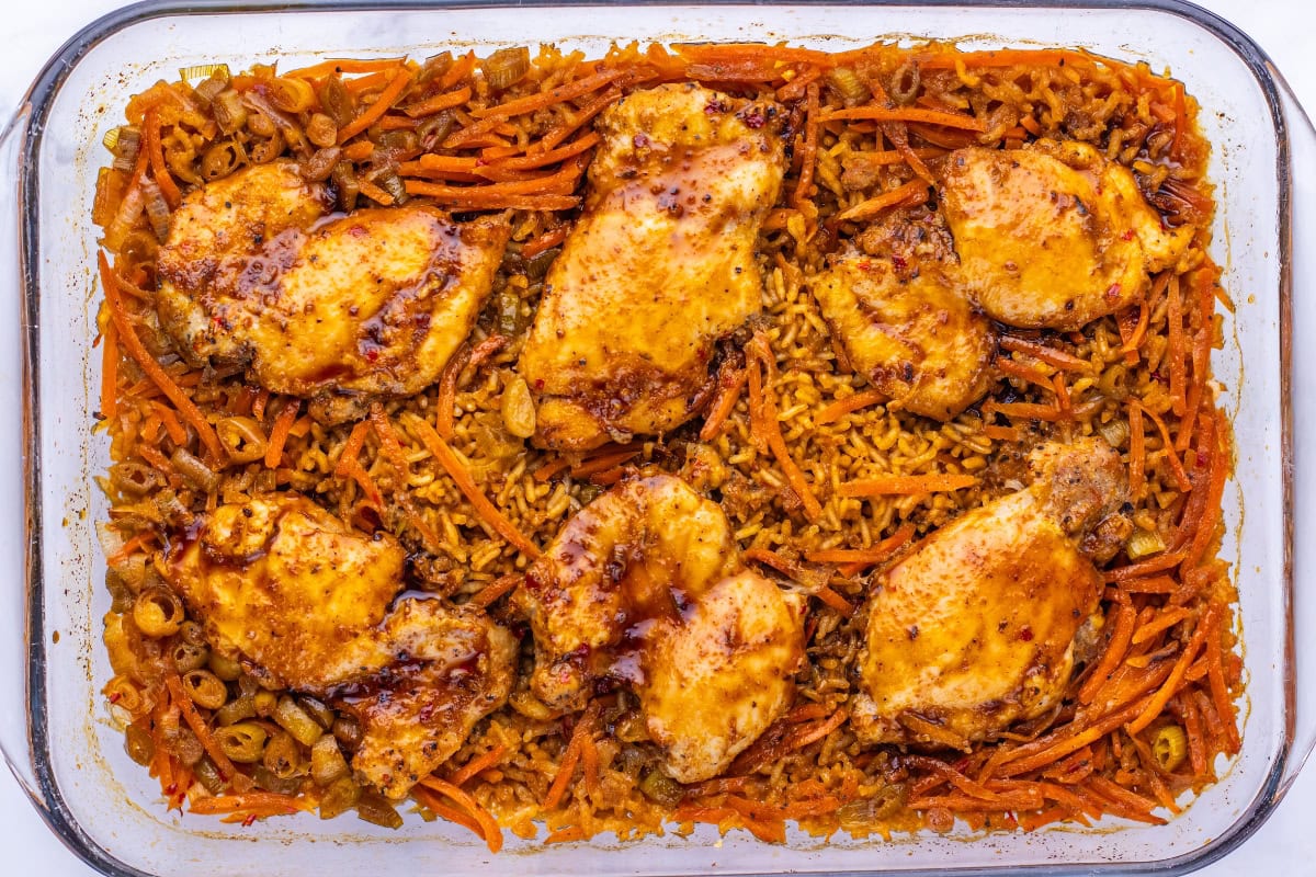 Baked chicken thighs with rice, shredded carrots, and olives in a glass dish.
