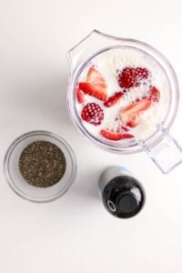 Blender with milk, strawberries, chia seeds, and a bottle on a white surface.