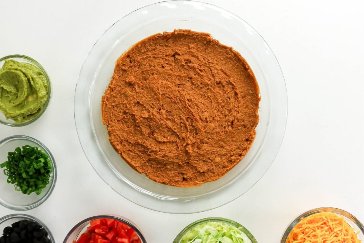 A pie dish with refried beans, surrounded by bowls of taco toppings.