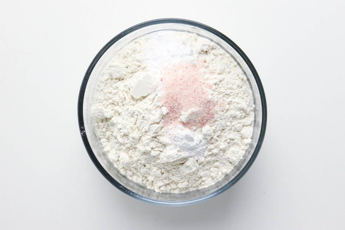 A glass bowl with flour, baking powder, and pink salt on a white surface.