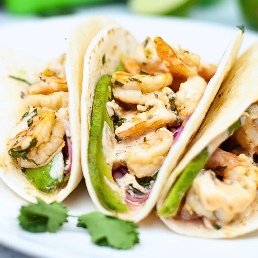 Three shrimp tacos with avocado slices and fresh herbs on a white plate.