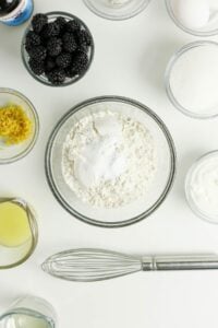 Baking ingredients in bowls on a white surface with a whisk at the bottom.