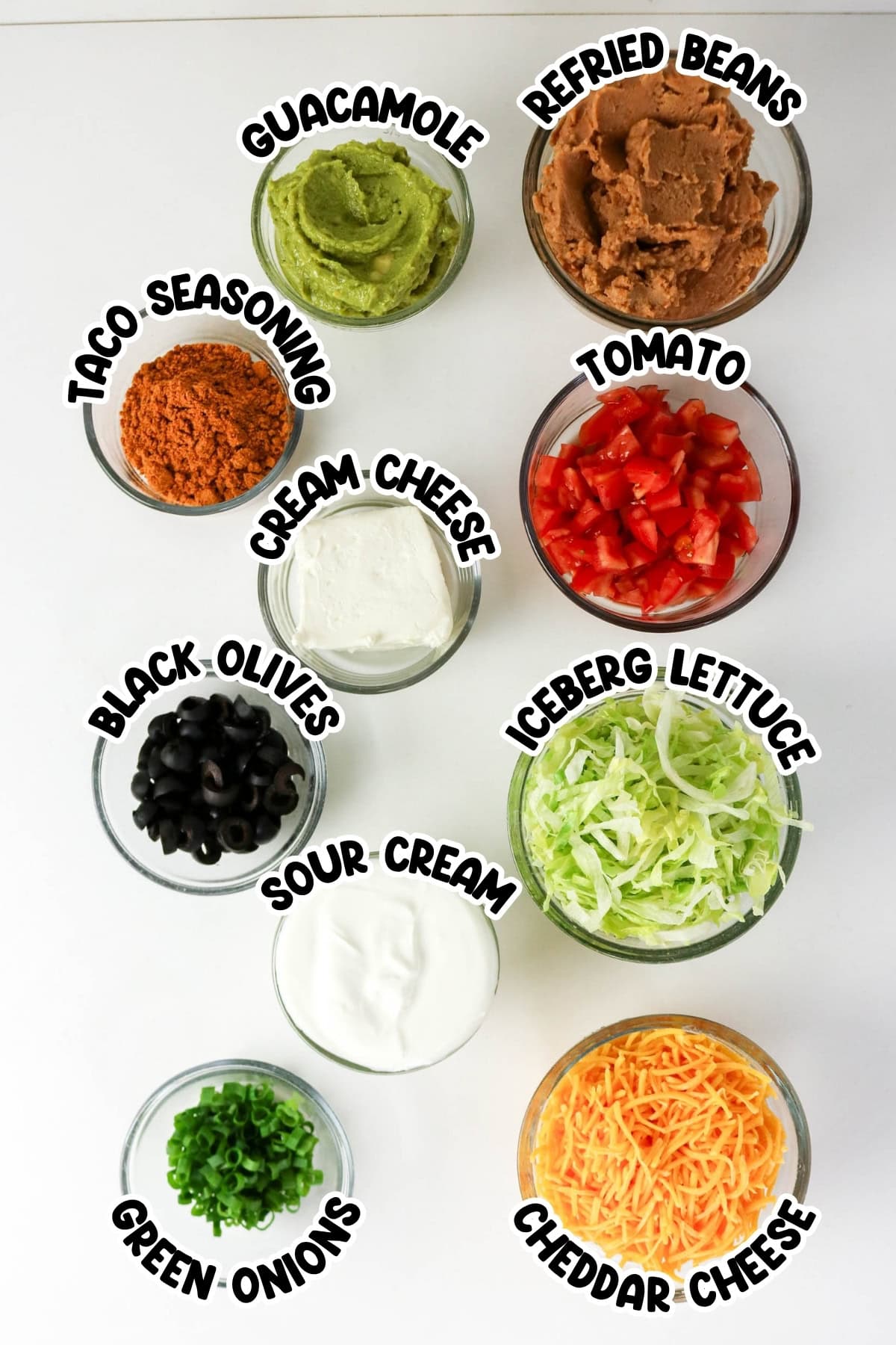 Nine bowls with taco dip ingredients labeled: guacamole, beans, cheese, and veggies.