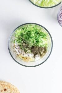 Glass bowl with yogurt, grated cucumber, herbs, and spices on a white surface.
